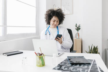 Arzt sitzt mit Laptop und benutzt ein Mobiltelefon in einer medizinischen Klinik - XLGF02782