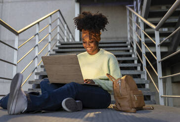 Happy young woman using laptop sitting on staircase - JCCMF05506