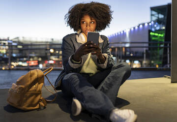 Junge Frau mit Handy in der Hand sitzt nachts auf dem Gehweg - JCCMF05511