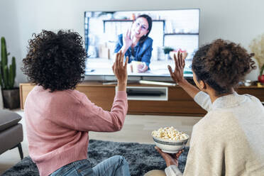 Frauen winken mit den Händen bei einem Videogespräch mit einem Freund über den Fernseher zu Hause - JSRF01878