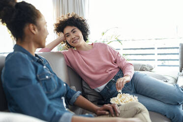 Lächelnde junge Frau mit Popcorn und Blick auf einen Freund, der auf dem Sofa im Wohnzimmer sitzt und spricht - JSRF01880