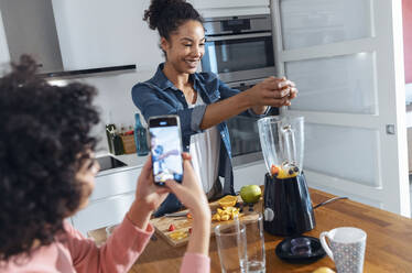 Woman photographing friend making juice in kitchen at home - JSRF01881