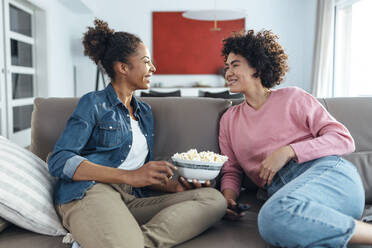 Glückliche Frau, die eine Schüssel Popcorn in der Hand hält und sich mit einem Freund auf dem Sofa zu Hause unterhält - JSRF01882