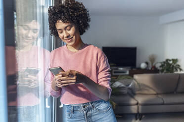 Smiling young woman using mobile phone leaning on glass window at home - JSRF01883