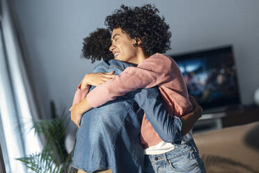 Happy woman embracing girlfriend standing in living room at home - JSRF01901