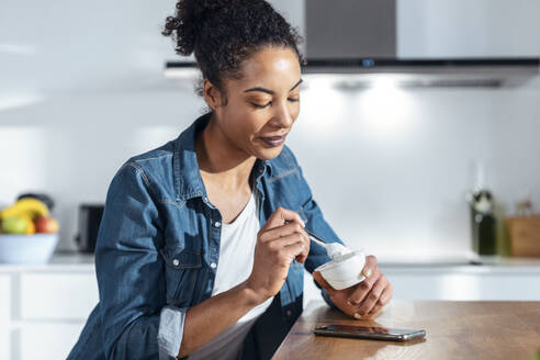 Frau isst Joghurt in der Küche sitzend zu Hause - JSRF01917