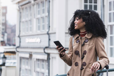 Smiling woman with curly hair holding mobile phone in city - PNAF03352