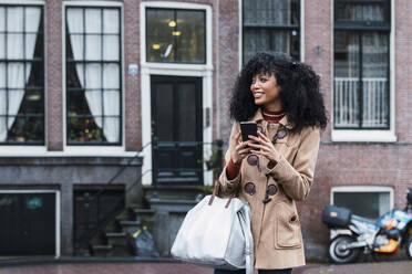 Happy young woman with purse holding smart phone in city - PNAF03357