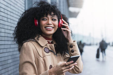 Glückliche junge Frau hält ihr Smartphone und hört Musik über drahtlose Kopfhörer - PNAF03359