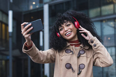Fröhliche Frau, die Musik über drahtlose Kopfhörer hört und ein Selfie mit ihrem Smartphone in der Stadt macht - PNAF03360