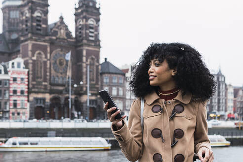 Lächelnde junge Frau mit lockigem Haar, die ein Mobiltelefon in der Stadt hält - PNAF03361