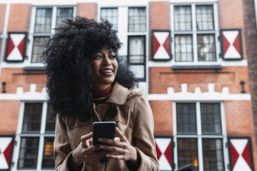 Happy woman holding mobile phone standing in front of building - PNAF03367