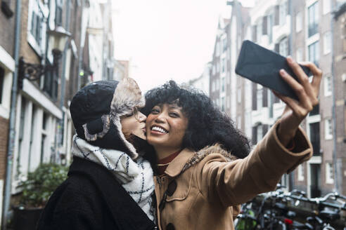 Glückliche junge Frau nimmt Selfie mit Freund küssen auf die Wange in der Stadt - PNAF03377