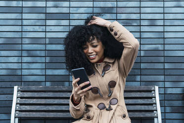 Lächelnde Frau mit Hand im Haar, die auf einer Bank sitzend ein Mobiltelefon benutzt - PNAF03381
