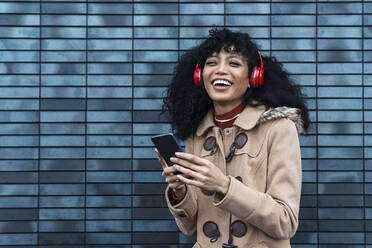 Glückliche Frau mit Mobiltelefon hört Musik über drahtlose Kopfhörer vor einer blauen Wand - PNAF03382