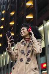 Lächelnde junge Frau mit Smartphone, die über drahtlose Kopfhörer in der Stadt Musik hört - PNAF03392