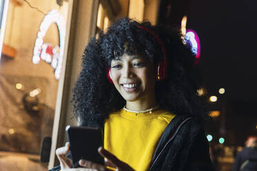 Glückliche junge Frau mit drahtlosen Kopfhörern, die nachts ein Mobiltelefon benutzt - PNAF03401