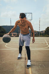 Junger tätowierter Latino-Junge spielt mit einem Basketball auf einem Platz - CAVF95399