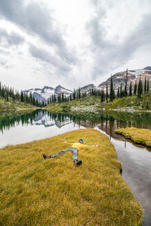 Young male hiker relaxes in long alpine grass by mountain lake - CAVF95498