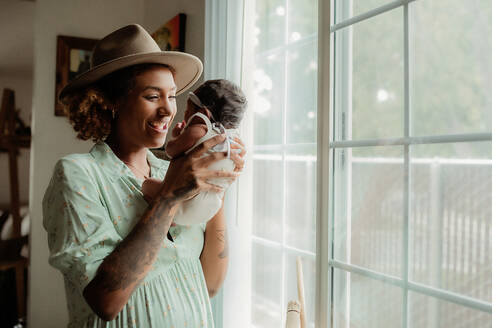 Lächelnde junge schwarze Mutter hält ihre kleine Tochter am Fenster - CAVF95503