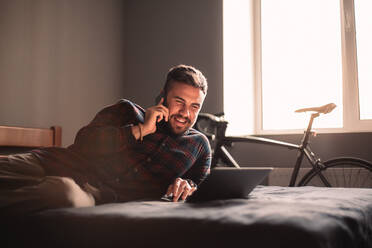 Happy man talking on smart phone while using laptop computer at home - CAVF95516