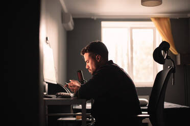 Man using smart phone and computer sitting at desk working at home - CAVF95518