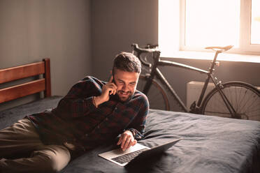 Happy businessman talking on smart phone using laptop computer at home - CAVF95528