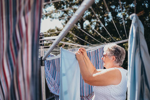Eine ältere Frau hängt Wäsche auf einer Wäscheleine im Freien auf. - CAVF95547