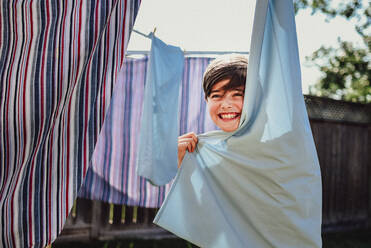 Happy boy hiding behind clothes hanging on a clothesline outside. - CAVF95548