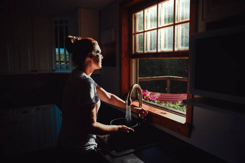 Woman filling a pot at the sink under a window in a dark kitchen. - CAVF95553