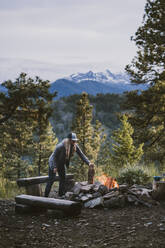 Woman wearing puffy jacket puts log onto campfire in the mountains - CAVF95593