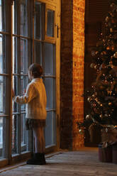 Ein Kind schaut am Weihnachtsabend aus dem Fenster, Seitenansicht. - CAVF95653