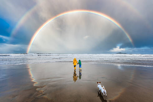 Hund läuft Kindern unter doppeltem Regenbogen am Strand hinterher - CAVF95663