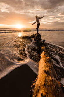 Teenager-Mädchen balanciert auf einem Baumstamm an einem Strand in Neuseeland bei Sonnenaufgang - CAVF95667
