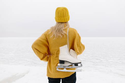 Woman in warm clothing standing with ice skates in front of winter sea - SIF00056