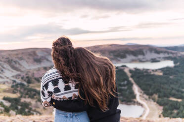 Rear view of two women hugging standing next to a lake with mountains - CAVF95692