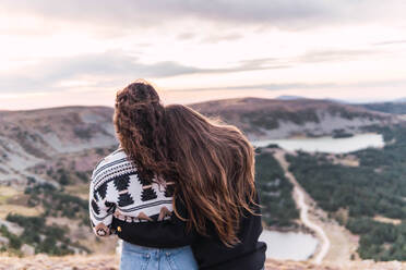 Rückansicht von zwei Frauen, die sich umarmen, neben einem See mit Bergen stehend - CAVF95692
