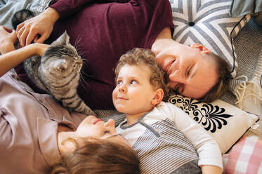 Eine dreiköpfige Familie liegt auf dem Bett und hat Spaß mit der Katze - CAVF95734