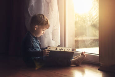 Side view of small boy sitting on the floor by window and readin - CAVF95751