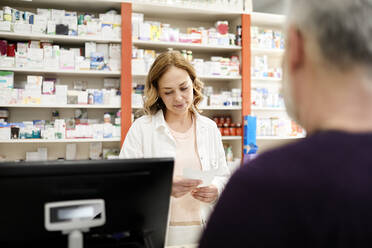 Female pharmacist reading prescription to customer at checkout counter in pharmacy store - ZEDF04430