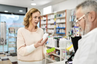 Customer holding bottle of medicine talking with pharmacist at checkout counter - ZEDF04474