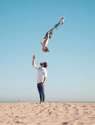 Full body side view of sporty female acrobat performing somersault high in air against blue sky over man standing on sandy seashore - ADSF33746