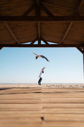 Seitenansicht eines entfernten, nicht erkennbaren Mannes, der eine Akrobatin in die Luft wirft, die einen Salto auf einem Sandstrand an einem Sommertag vollführt - ADSF33750