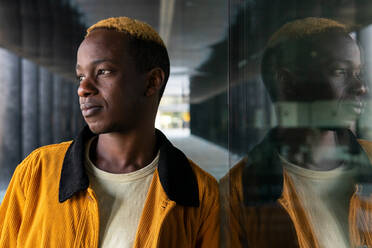 Confident African American male with dyed hair looking into distance while standing near modern building with glass wall in city - ADSF33754