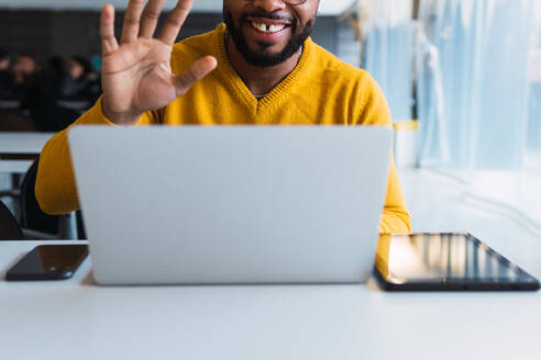 Abgeschnittene unerkennbare fröhliche afroamerikanische bärtige Mann mit Zahnlücke Unternehmer in Pullover und Brille mit Video-Anruf auf Netbook mit Kopfhörer bei der Arbeit im Büro - ADSF33755