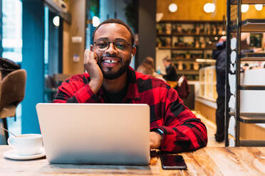 Positiver afroamerikanischer, bärtiger, männlicher Freiberufler, der mit Kopfhörern auf seinem Netbook surft, während er in einem Café arbeitet und in die Kamera schaut - ADSF33757