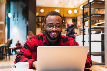 Positive African American bearded male freelancer browsing netbook with earphones while working remotely in cafe and looking at screen - ADSF33758