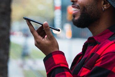 Fröhlicher, unkenntlich geschnittener, bärtiger Afroamerikaner in kariertem Hemd und Hut, der eine Sprachnachricht auf einem Mobiltelefon in einer städtischen Straße aufnimmt - ADSF33762