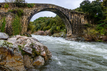 Schäbige Steinbogenbrücke über einen reißenden Fluss, der zwischen felsigen Küsten mit üppig grünen Bäumen in der Natur an einem Sommertag in Extremadura, Spanien, fließt - ADSF33763