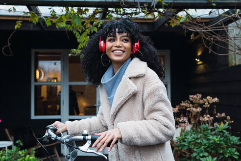 Side view smiling young African American female with curly dark hair wearing warm coat walking with bicycle while listening to music in headphones near house in autumn day - ADSF33774